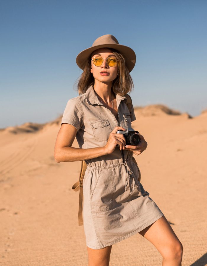 stylish young woman in khaki dress walking in desert, traveling in Africa on safari, wearing hat and backpack, taking photo on vintage camera, exploring nature, hot summer day, sunny weather